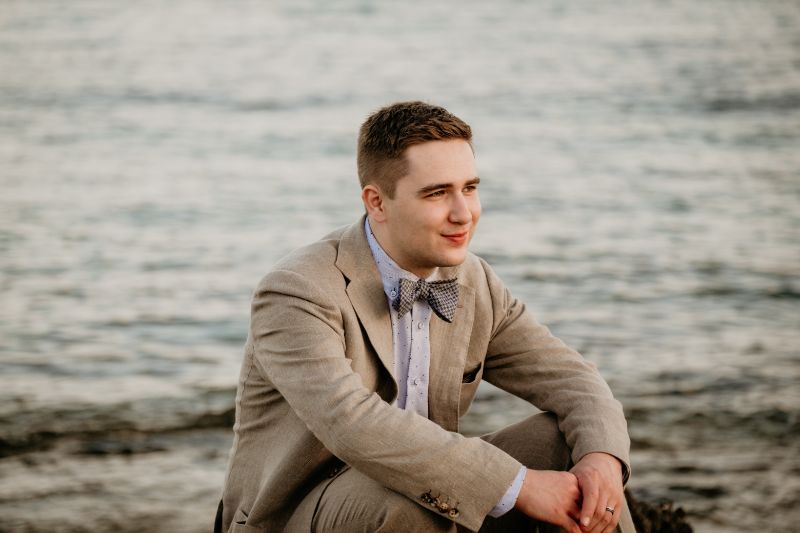 A young man sitting on a beach in a beige linen suit