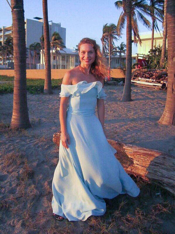 A young woman on a beach with palm dress in the background, wearing a white wedding dress and smiling